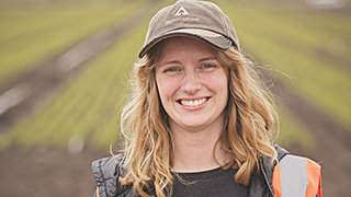 Smiling woman wearing Ƶ baseball cap in foreground of field of newly planted trees.